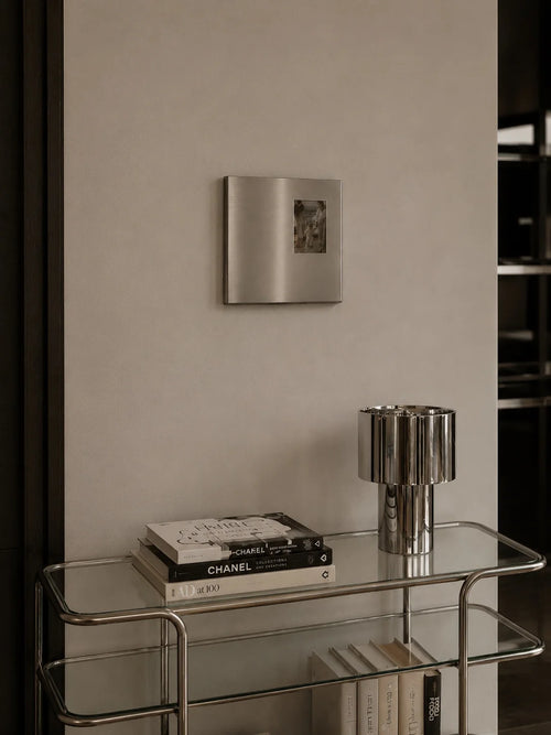 Ferrani Mono Frame on a beige wall above a glass console with a chrome lamp and stacked books.