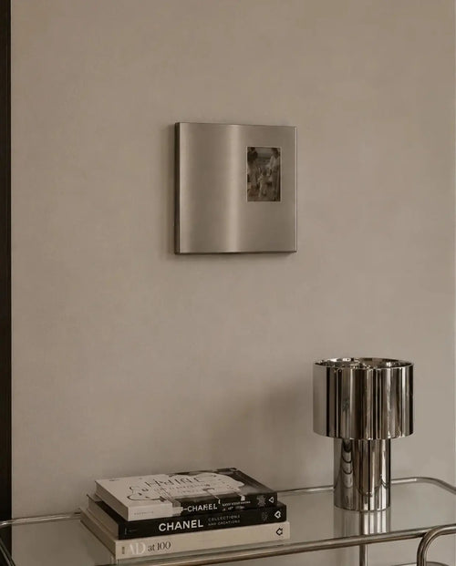 Ferrani Mono Frame on a beige wall above a glass console with a chrome lamp and stacked books.