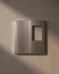 Empty Ferrani Mono Frame in brushed stainless steel on an exposed concrete wall, with light passing through the opening.