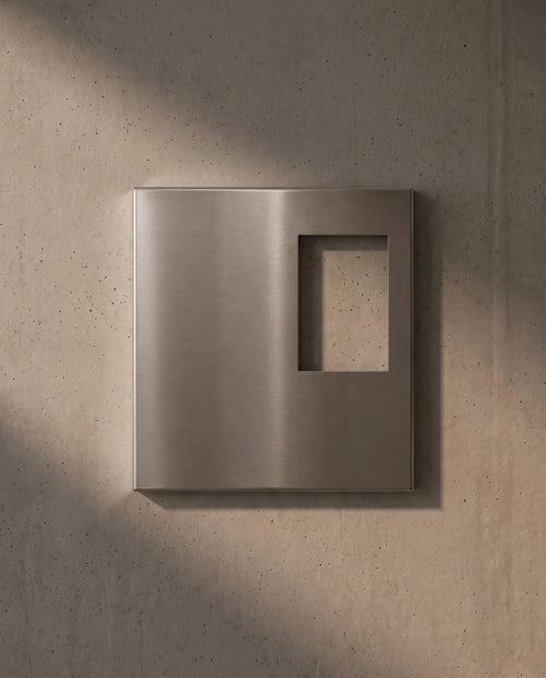 Empty Ferrani Mono Frame in brushed stainless steel on an exposed concrete wall, with light passing through the opening.